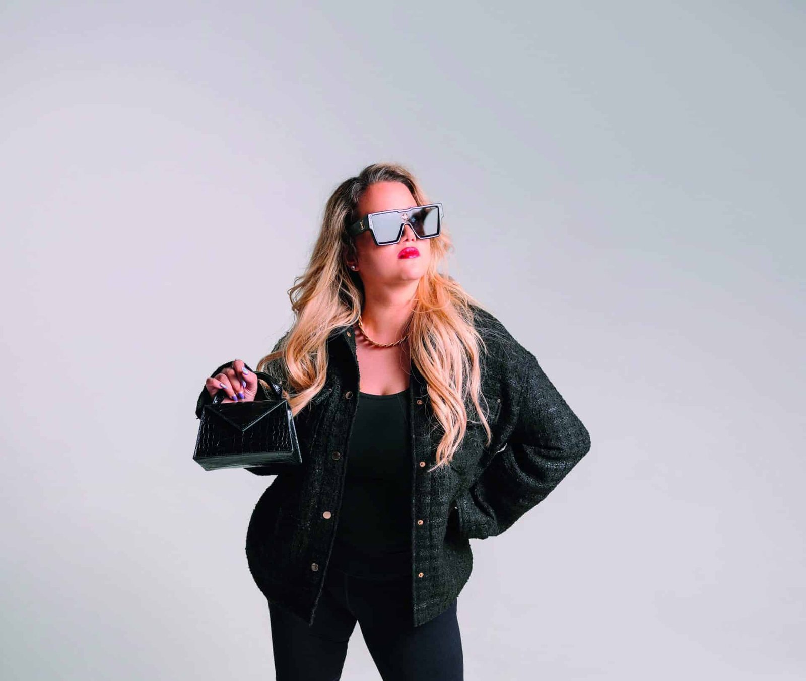 A fashion-forward woman in a black jacket and sunglasses holding a handbag.