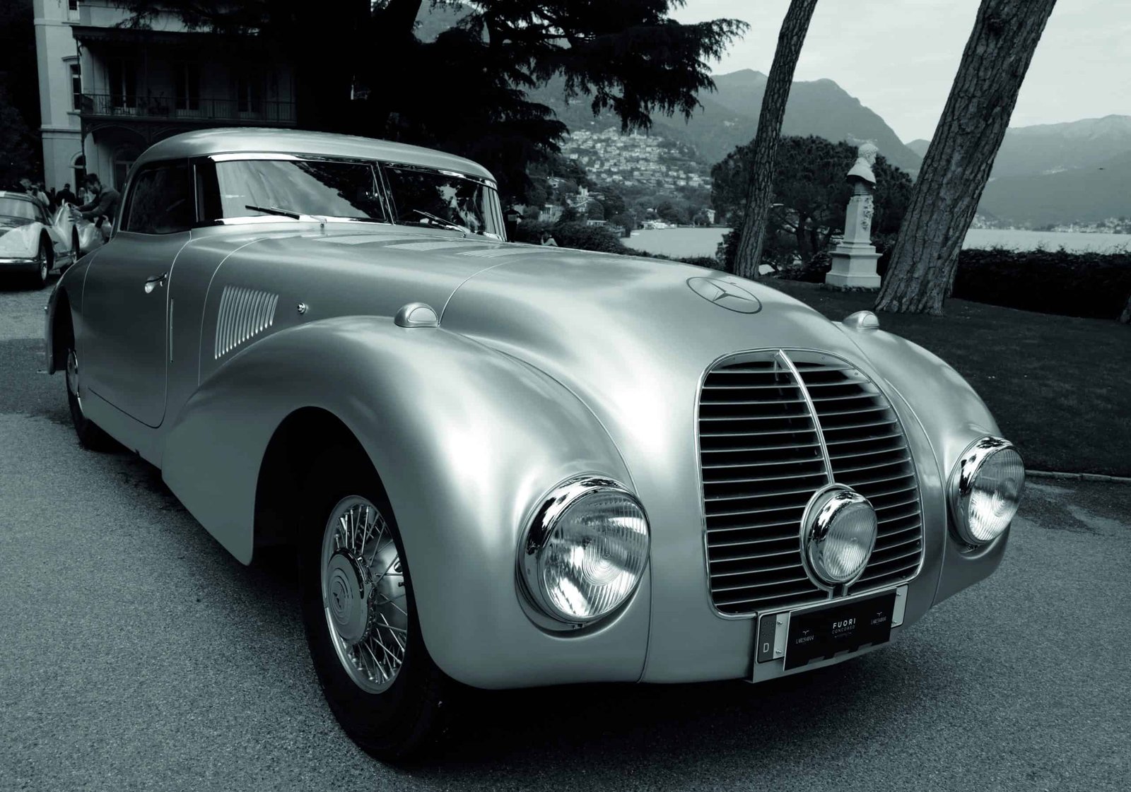 A silver vintage car is parked in front of a wind-shaped lake.
