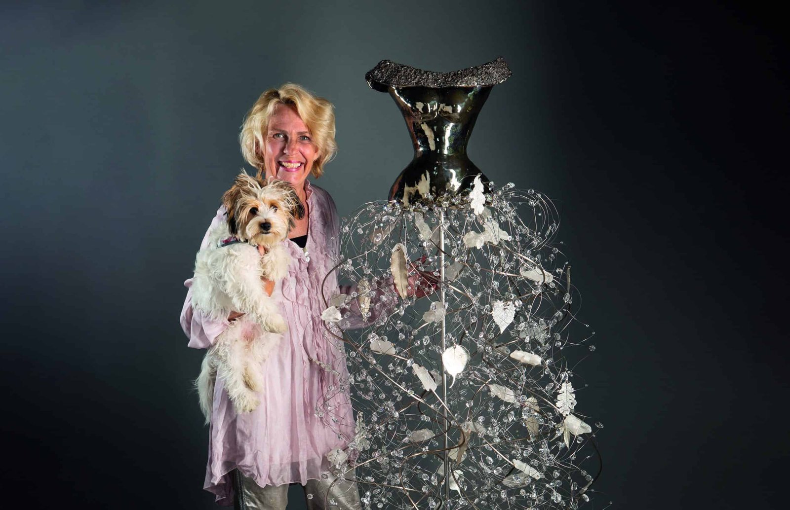 A feminine woman holds a dog in front of a large sculpture.