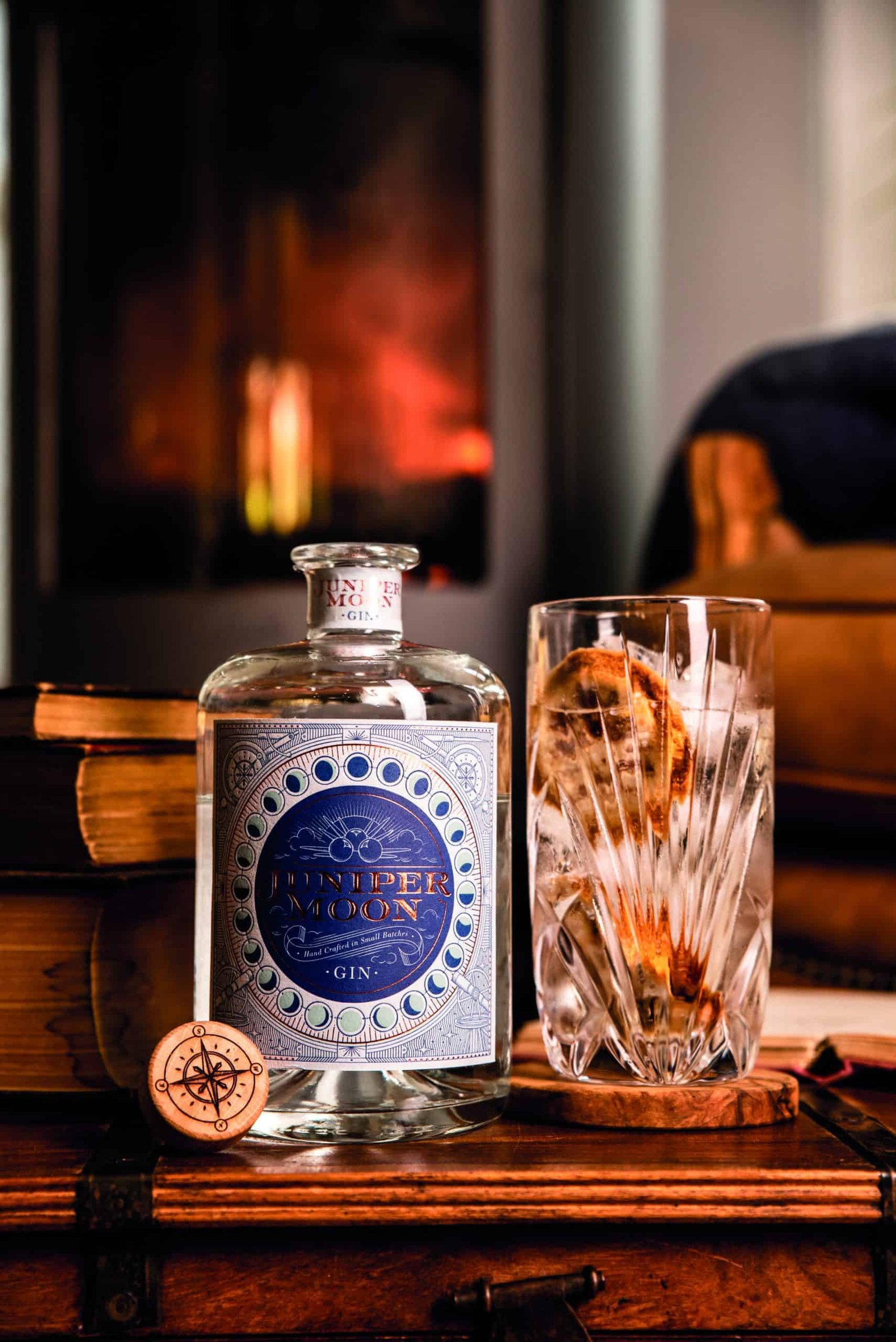 A glass of gin and a book on a table next to a fireplace that sets the mood for a peaceful journey into the depths of imagination.