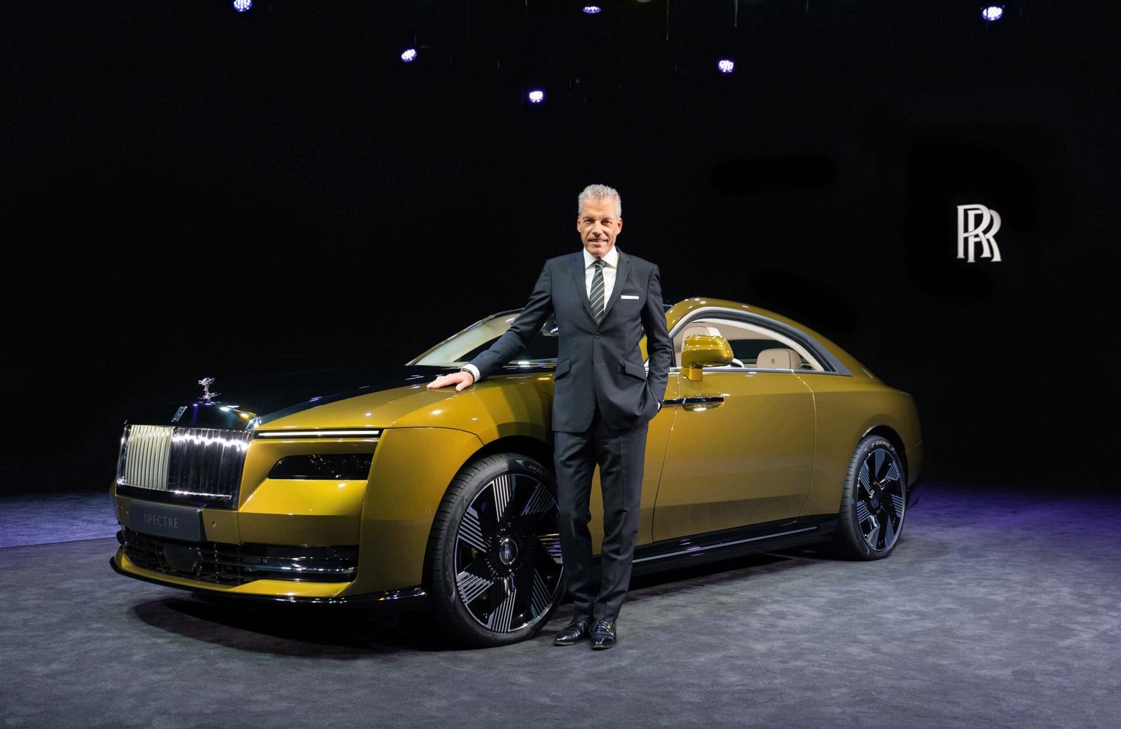 A bold man in a suit standing next to a Rolls Royce, embodying the spirit of innovation.