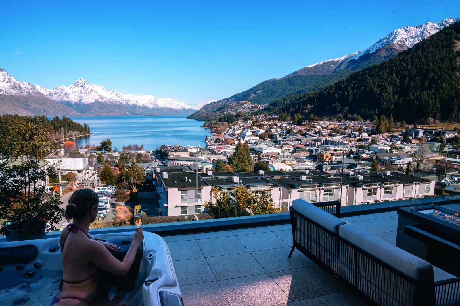 A woman sitting in a hot tub on a penthouse balcony overlooking a lake and mountains.